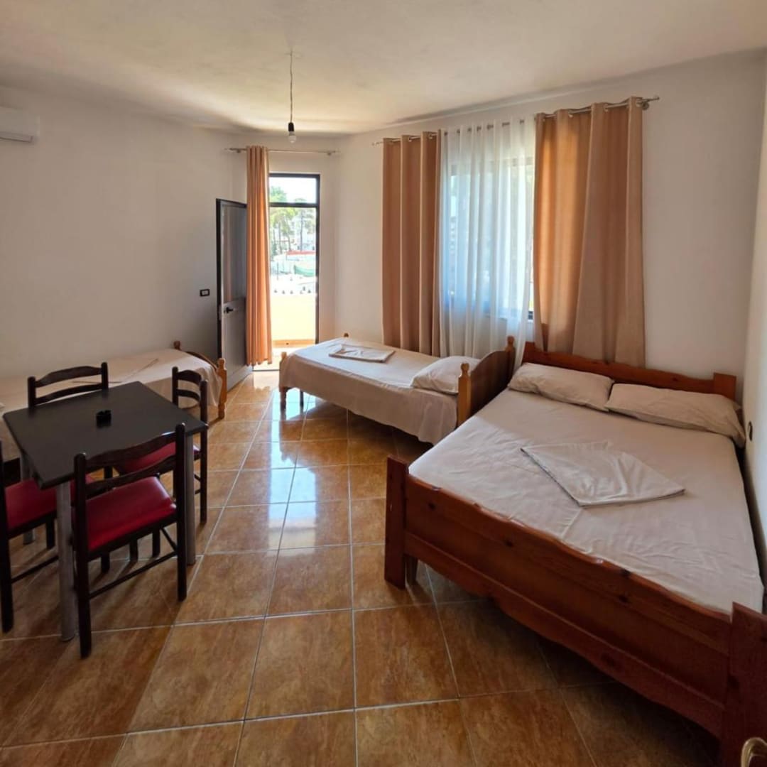A double bed, two single beds and a table with red chairs in a tiled room, with the balcony door at the far end