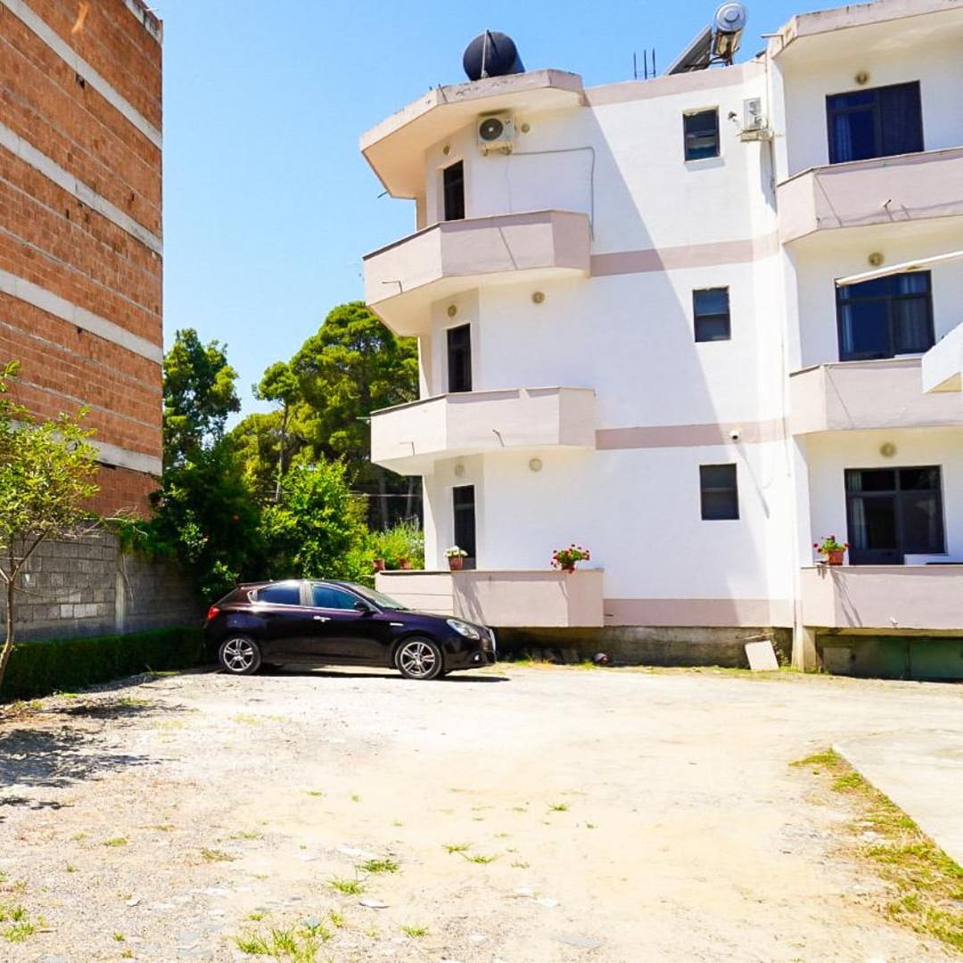 A dark car parked on a gravel yard beside a white building with balconies, with an unfinished brick building behind a block wall