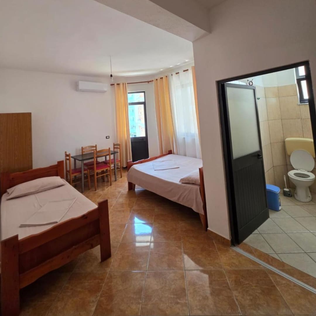 A single bed and a double bed in a tiled studio, with a table and four chairs by the balcony door and the bathroom door open