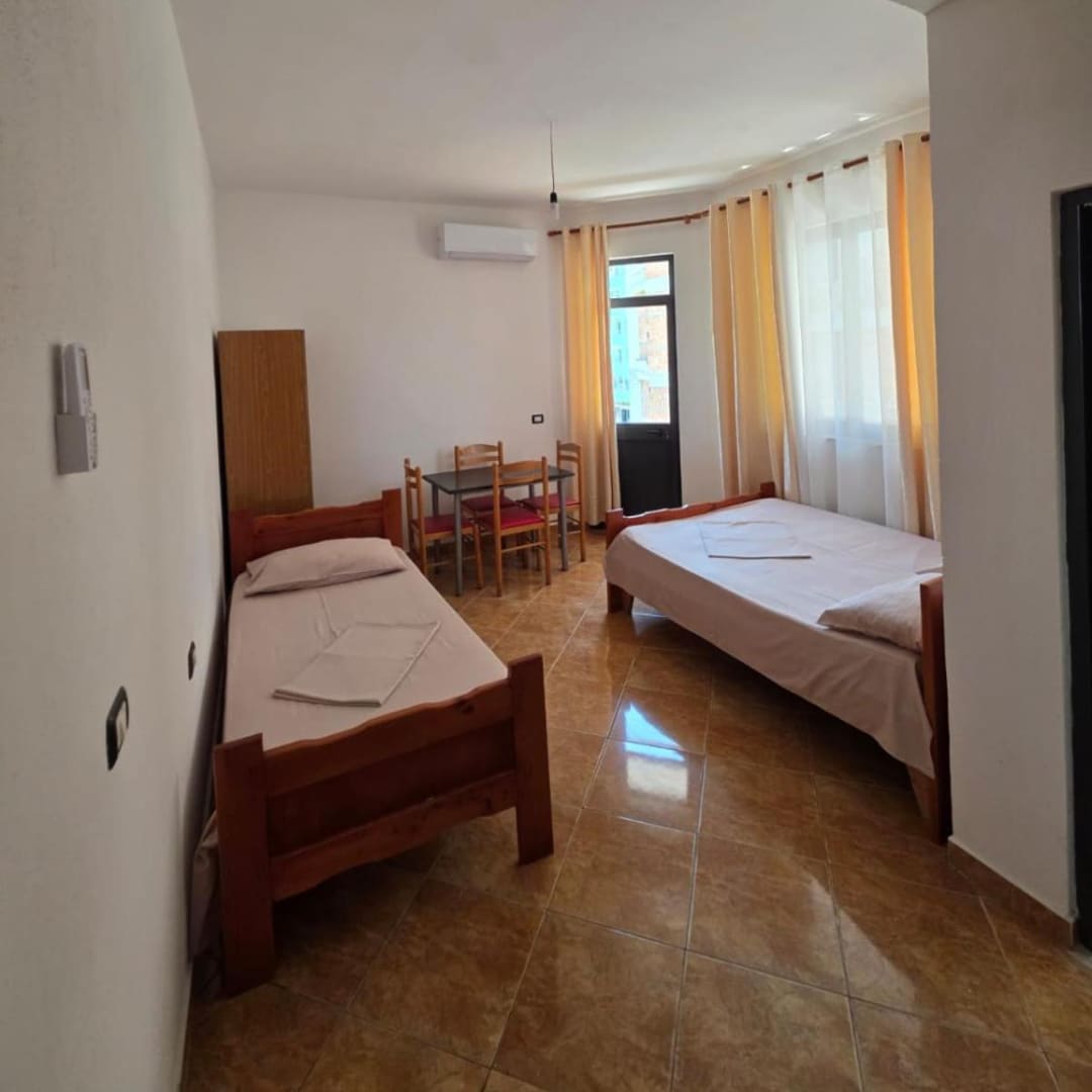 A single bed and a double bed in a tiled studio, with a wardrobe, a table and four chairs, and curtained windows by the balcony door
